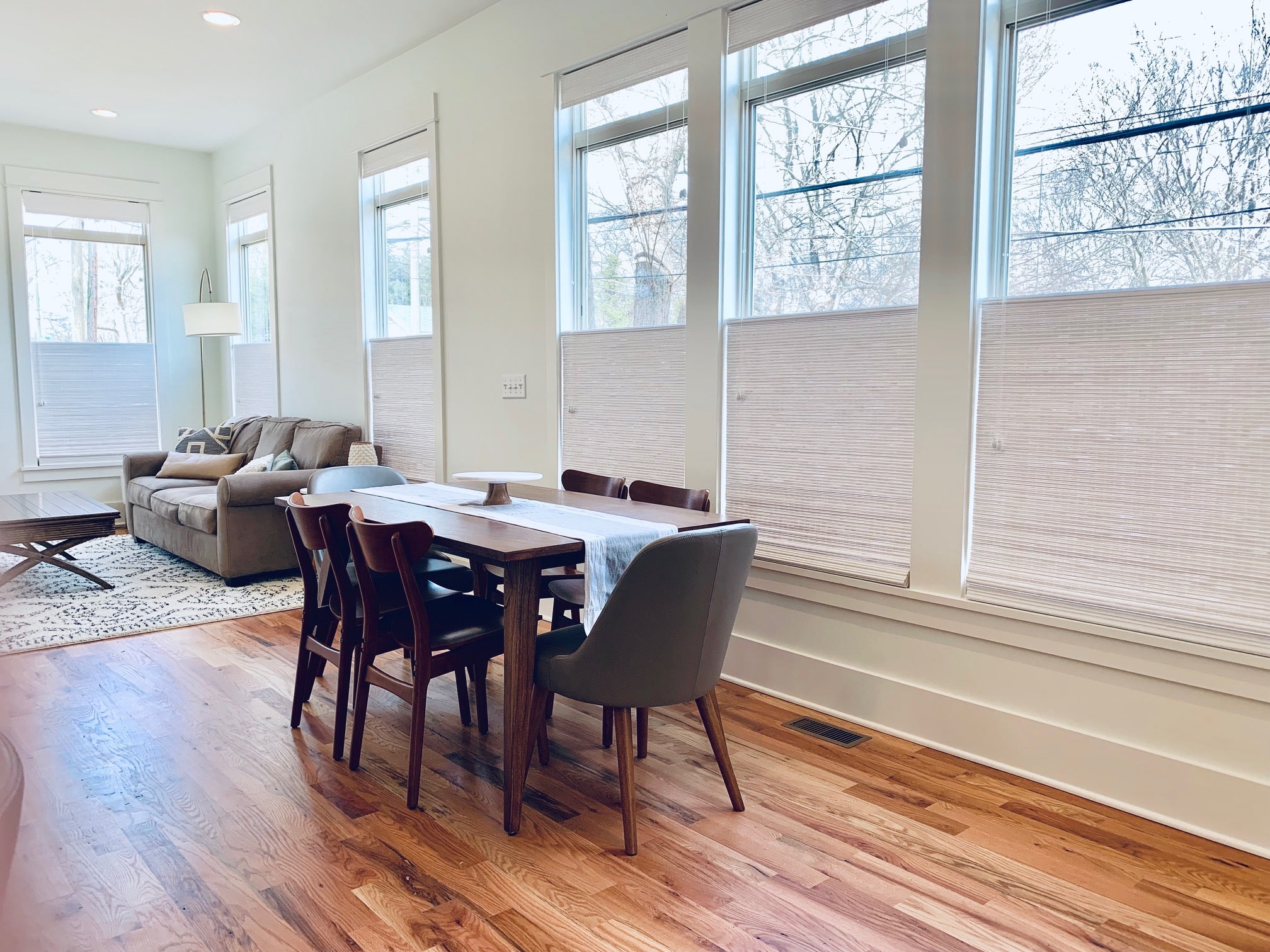 Top down woven shades partially cover six large windows in an open dining room and den area