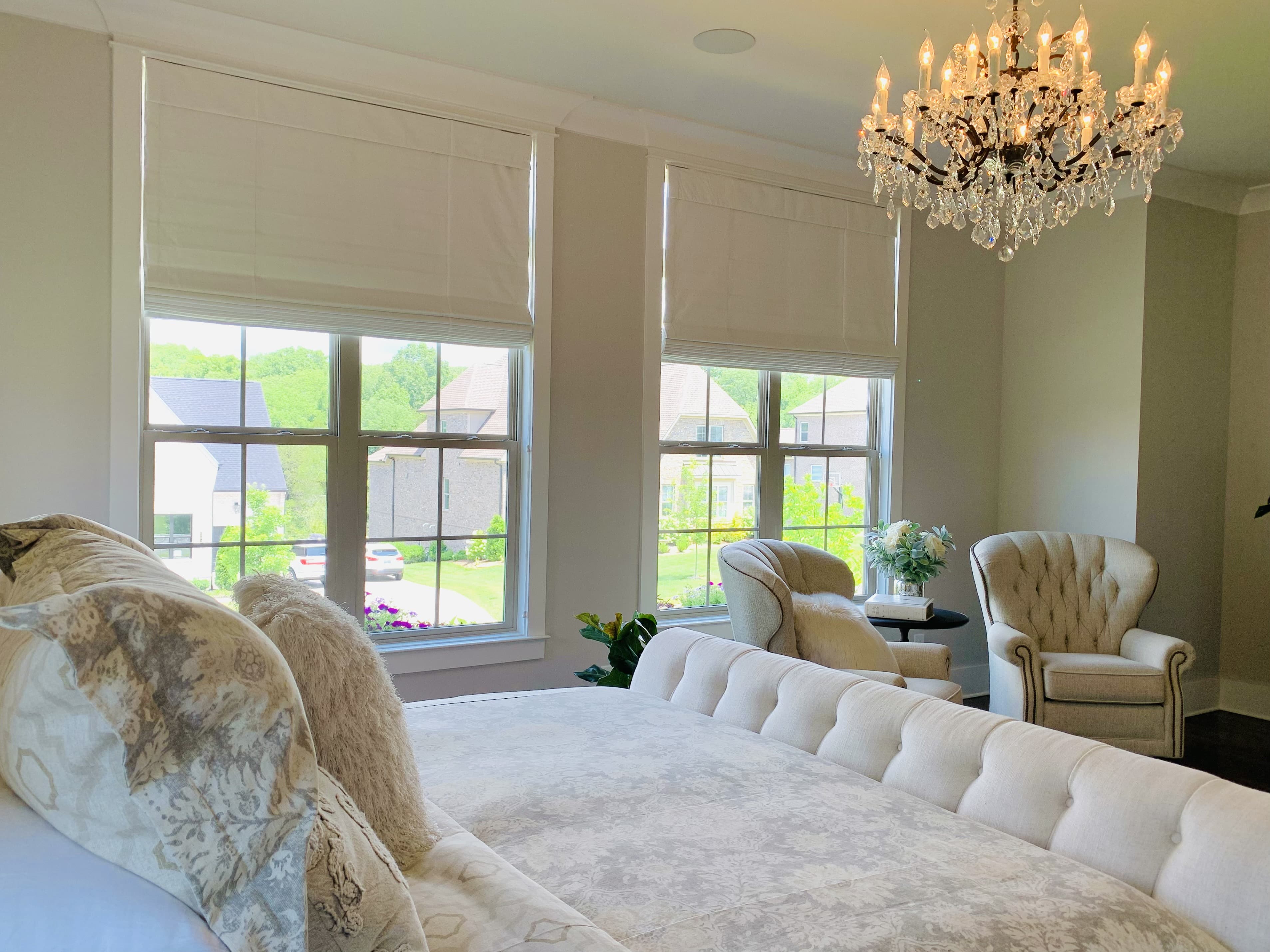 Two roman shades halfway lowered on two windows behind a bed beside two chairs