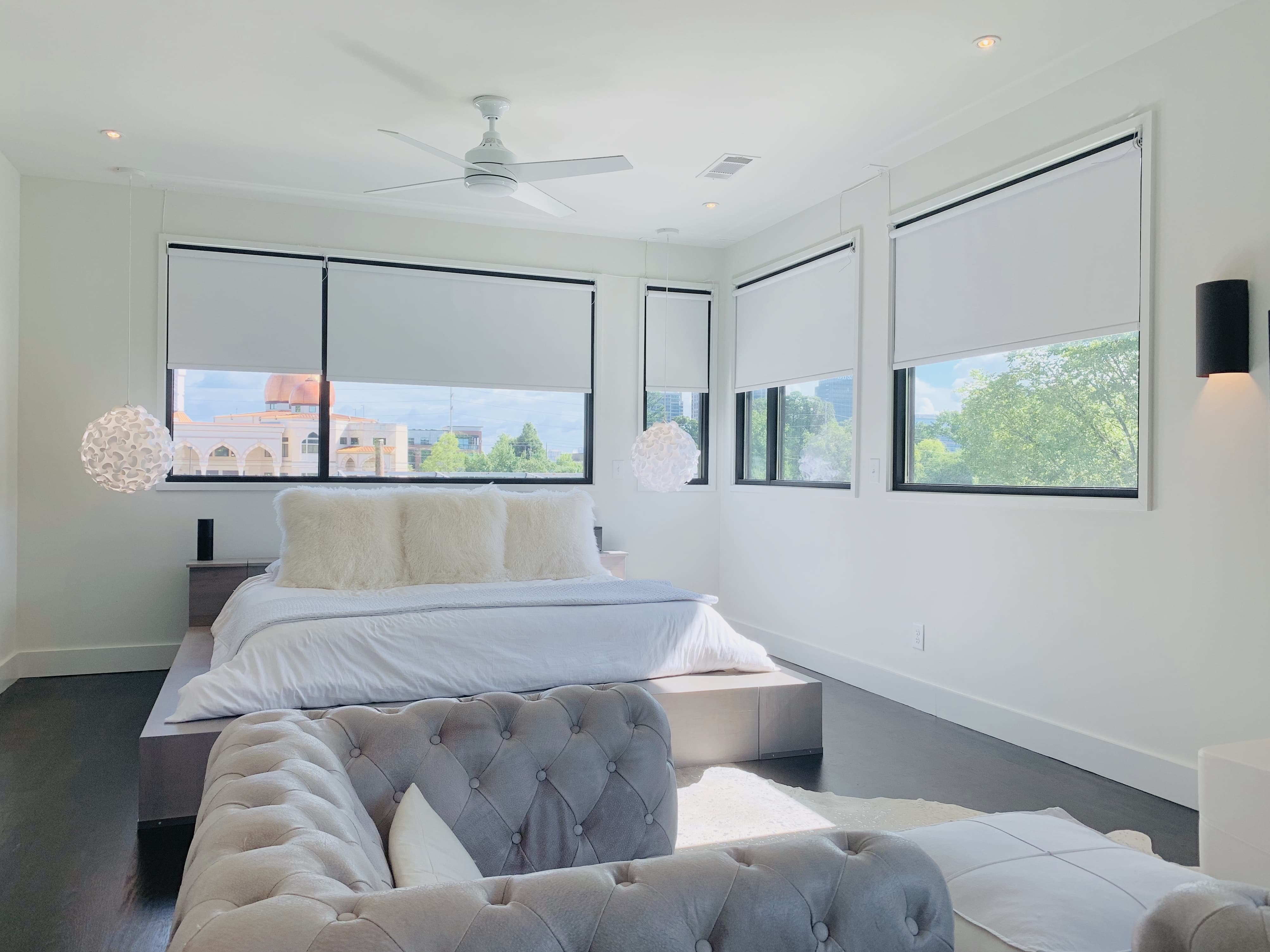 Five white roller shades lowered halfway on large windows behind a bed and large armchair