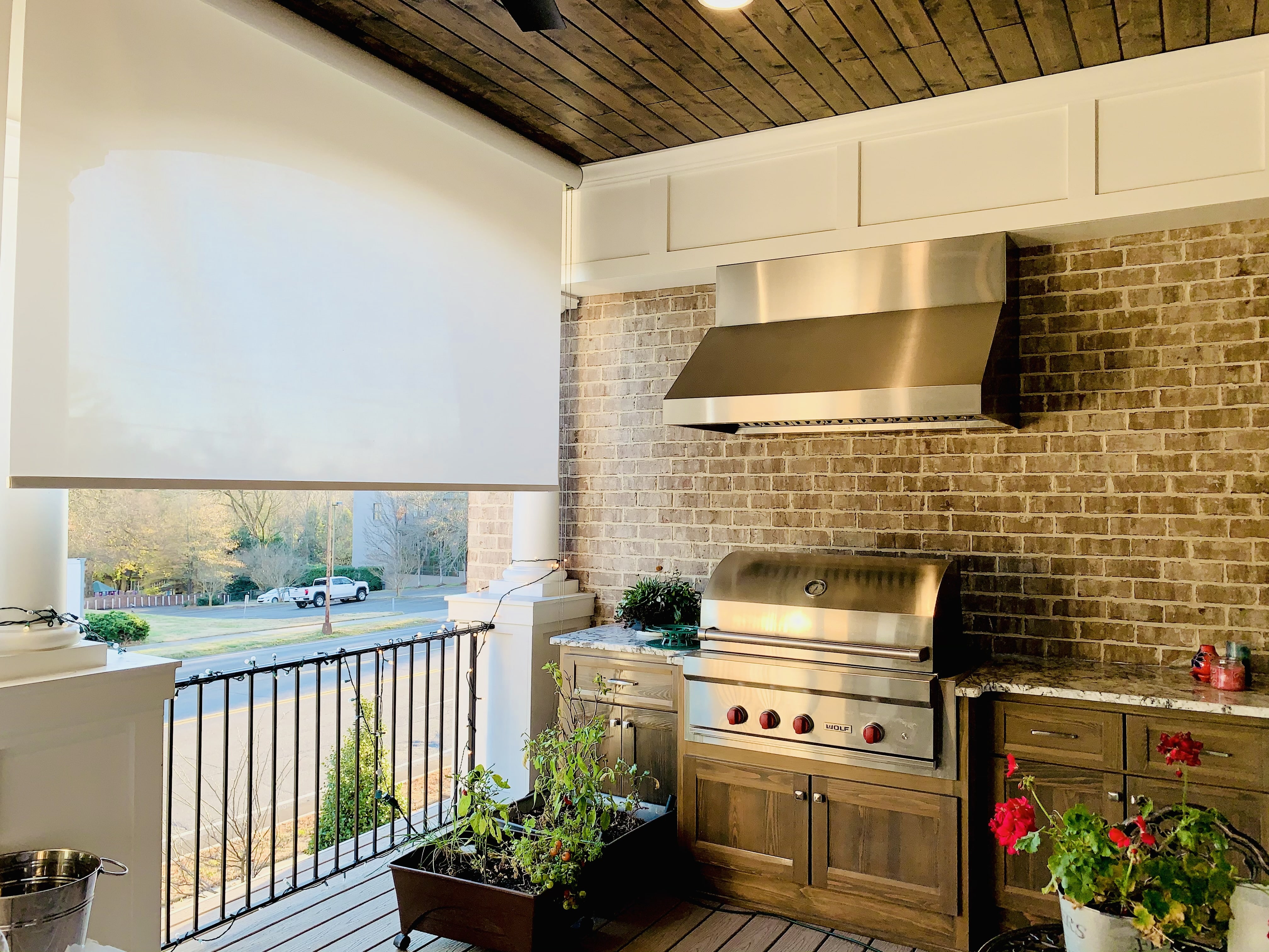 A large white roller shade diffuses light onto a porch with a grill and potted plants