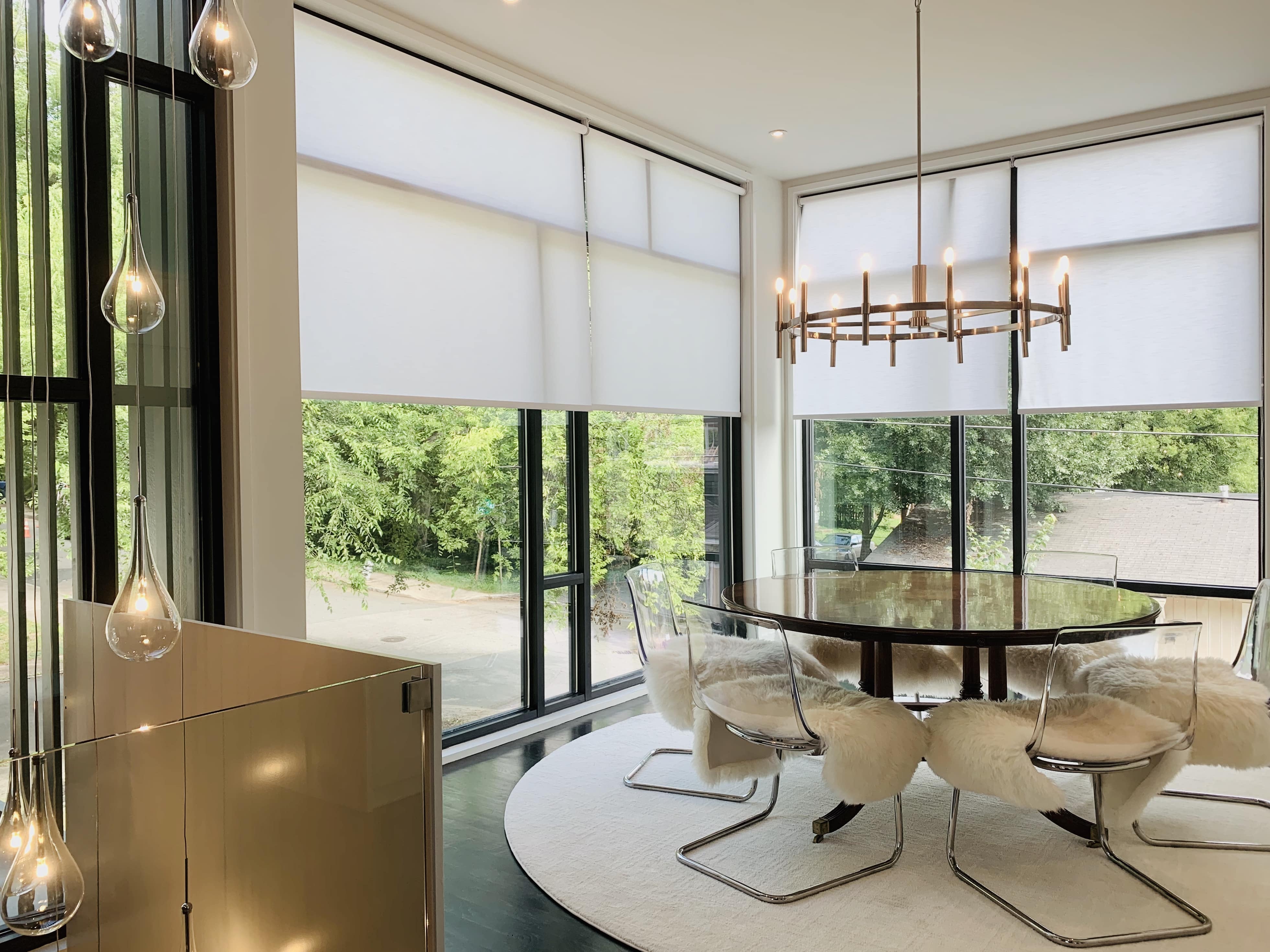 Two white roller shades lowered halway down two large windows in a dining room with a glass table