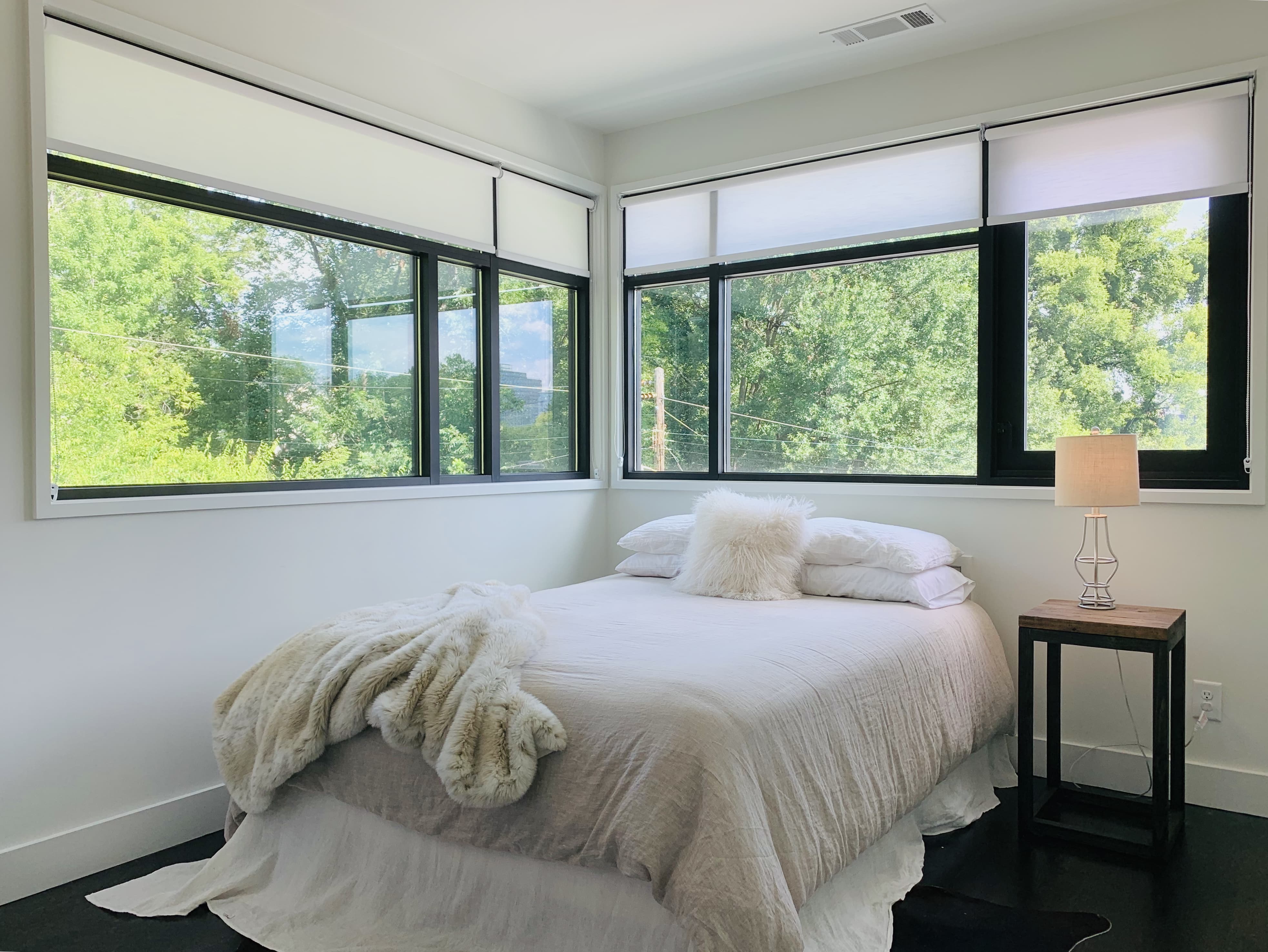 Four roller shades partially cover large windows behind a bed with a bedside table