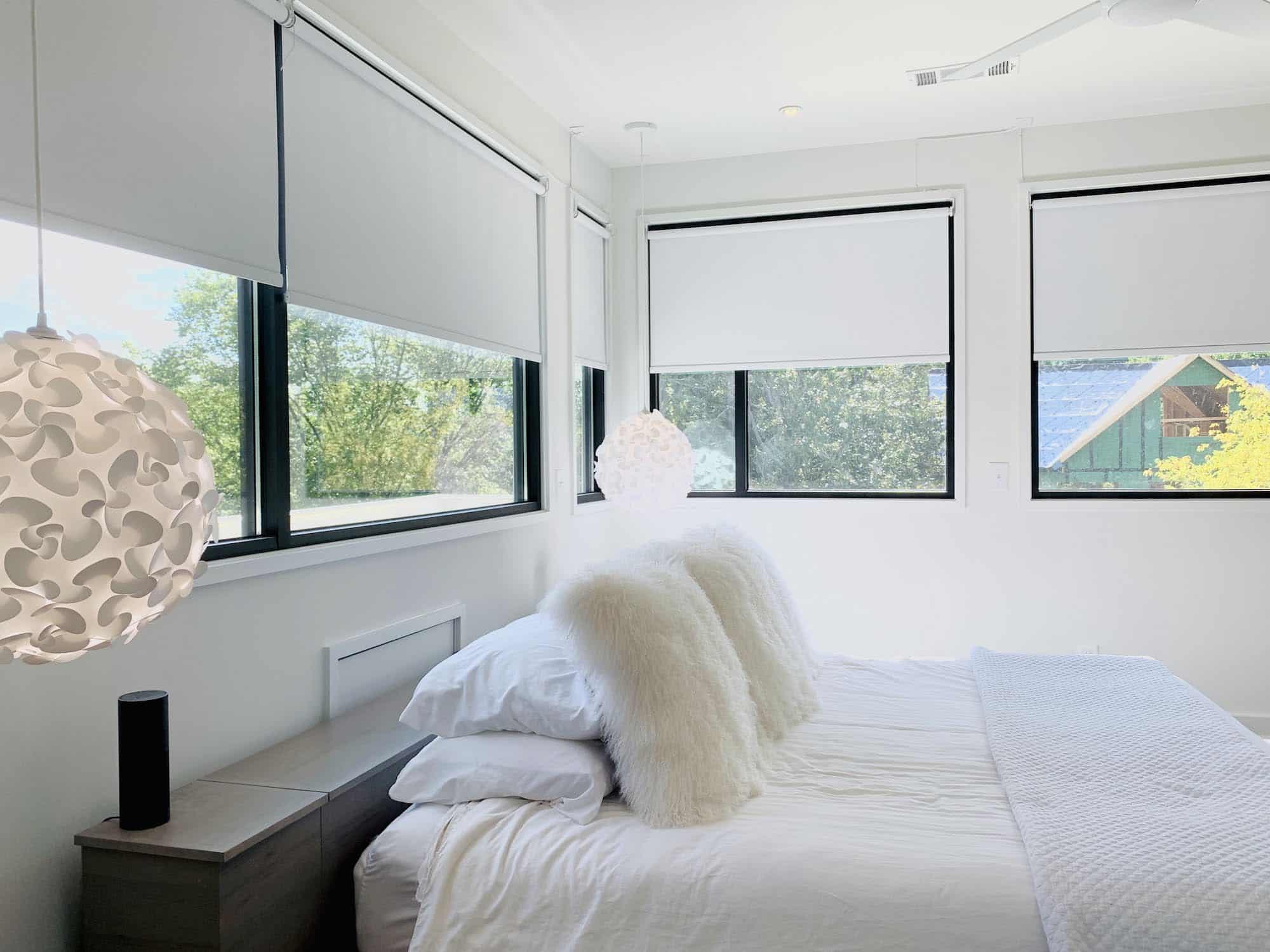 A white bedroom with five large windows, on which white roller shades are rolled up halfway